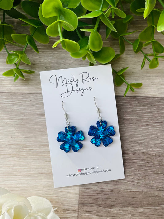 Photograph of one pair of epoxy resin earrings in dark royal blue glitter mini flower shape on timber background.