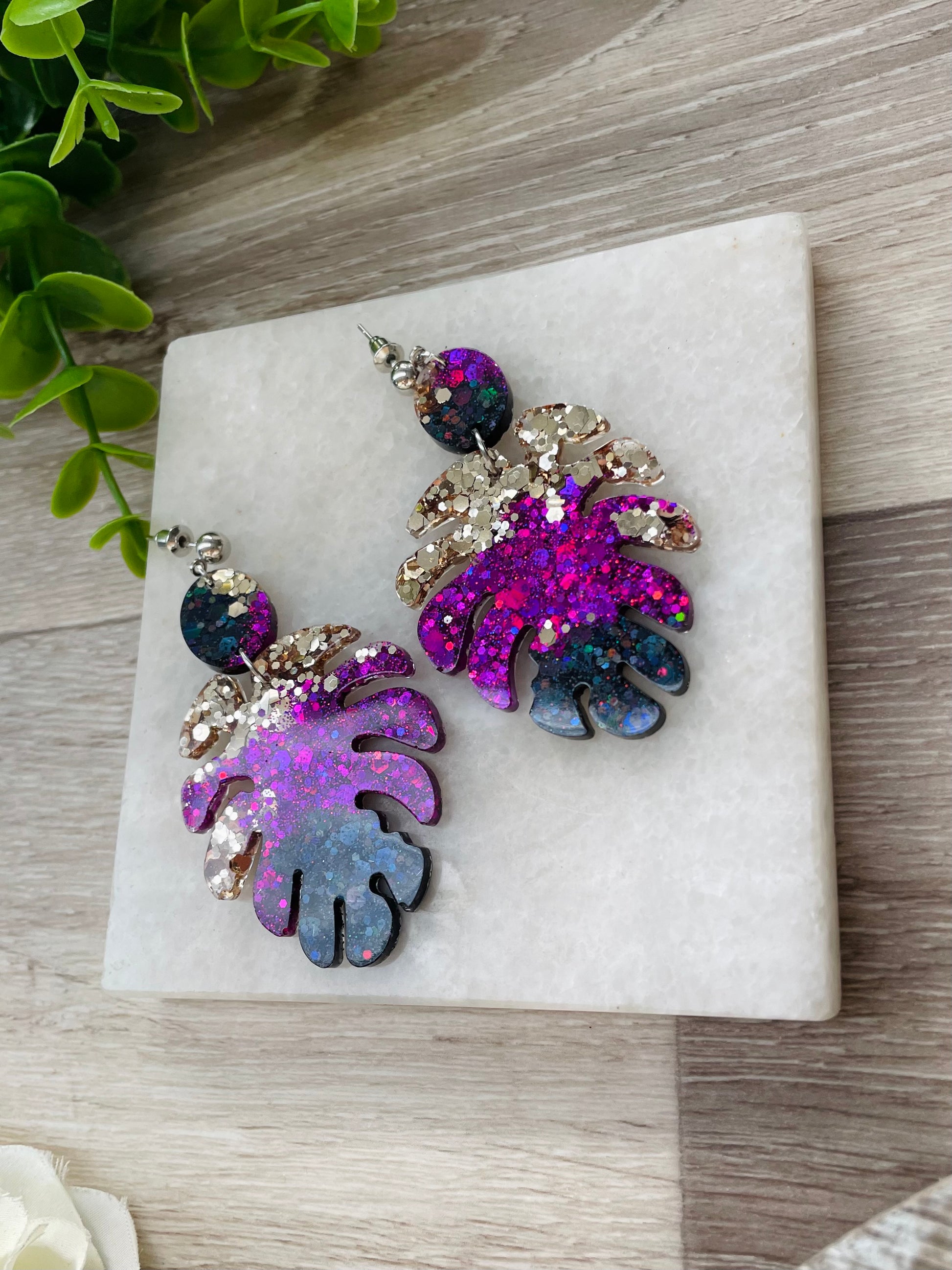 Photograph of one pair of epoxy resin earrings in monstera shape on timber background. The colours of these earrings are black, vivid purple & light gold.  
